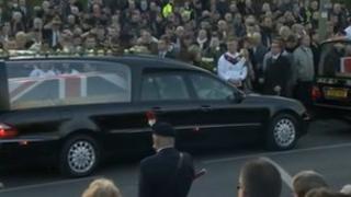 Two hearses drive past a large crowd in Carterton
