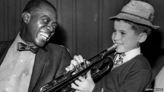 Louis Armstrong smiles as he hears Enrico Tomasso, nine, from Leeds play the trumpet in 1970