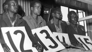 14 April 1953: Suspected members of the Mau Mau, accused of murder on trial. They are sitting in a shed holding large identifying numbers on their laps