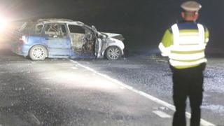 Police officer standing near car which has been destroyed by fire