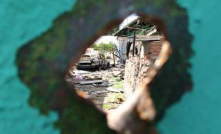 View through a hole in a wall caused by shrapnel shows destroyed building and burned out car in Marakove