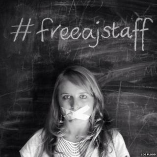 A journalist with taped up mouth against a blackbard with #FreeAJStaff