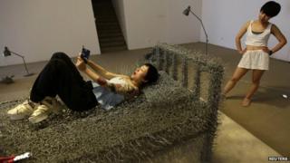 A woman watches as another takes a selfie on a iron wire bed at Beijing Art Now Gallery.