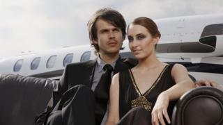A young man and woman sit on a couch in front of a private jet.