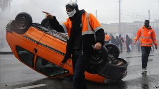 The Peugeot car lying on its roof with a protestor next to it