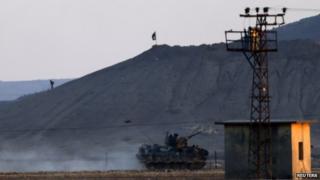 An Islamic State fighter walks near the group's black flag on a hill near the besieged Syrian town of Kobane as a Turkish military vehicle passes just across the border, 7 October