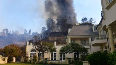 A huge plume of black smoke rises from a burning home on a hilltop beside one still standing in Bel Air, east of the 405 freeway on December 6, 2017 in Los Angeles, California