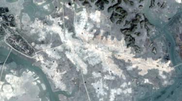 A satellite image shows the layout of a village in bright white - apparently bulldozed ground - in stark contrast to the green landscape around it