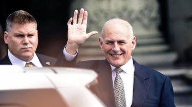 John Kelly waves to a camera over the roof of his car, flanked by a man who appears to be a security guard