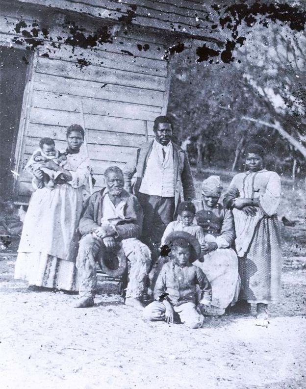 Fotografia de famÃ­lia de escravos nos Estados Unidos - mulher com crianÃ§a no colo, senhor sentado, homem adulto em pÃ©, duas mulheres e duas crianÃ§as