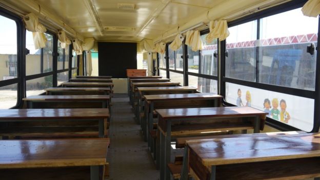 Interior de uma sala-ônibus, ônibus reformado para ser sala de aula; ao fundo um quadro-negro, à frente, carteiras