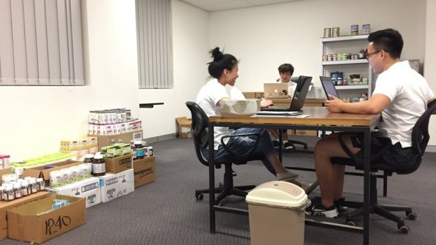 Three workers sit in an office surrounded by products to the shipped to China