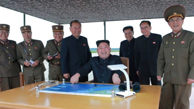 Kim Jong-un, seated in centre at a desk with a map, is seen surrounded by smiling officials and military personnel in an official photo