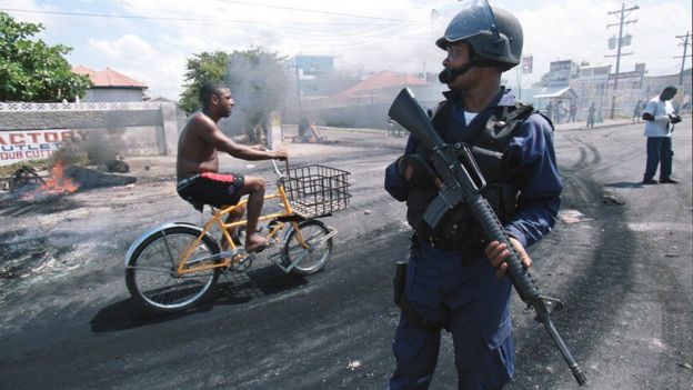 Policía sostiene un arma mientras un hombre pasa en bicicleta detrás de él. En el fondo hay neumáticos quemados.