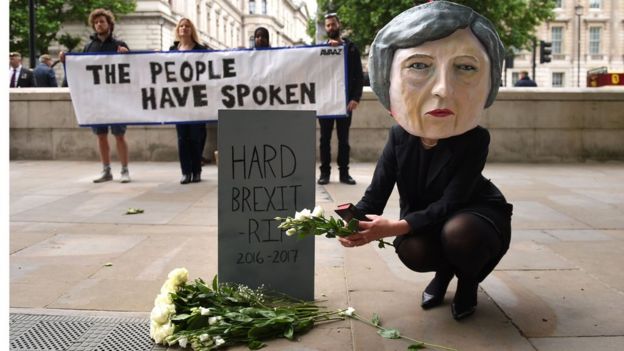 Alife-size giant-headed puppet of Theresa May leaving flowers at a tombstone bearing the words Hard Brexit RIP
