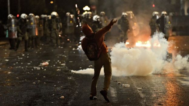 A hooded protester throws a rock at a line of riot police in Athens. A canister of tear gas is deployed on the street.