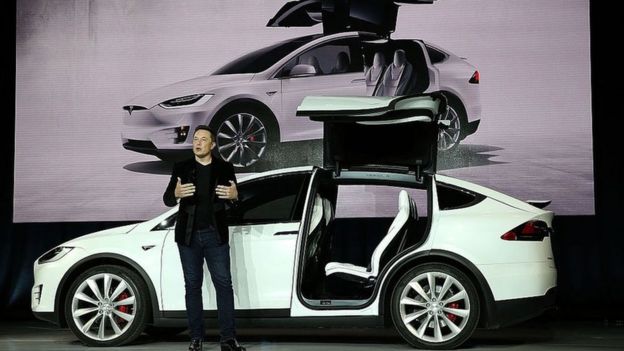 Elon Musk da una conferencia con un Tesla detrás