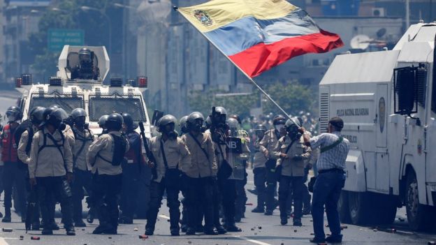 Un hombre ondea la bandera de Venezuela ante un grupo de guardias nacionales con equipos antimotines.