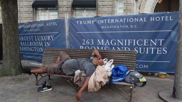 Un hombre sin hogar duerme en un banco en una calle frente a un hotel de lujo.