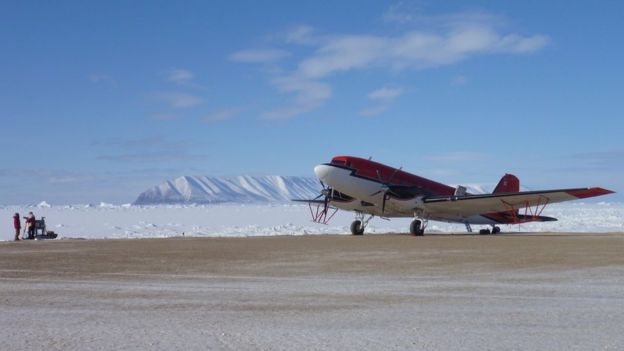 A red aeroplane stationary on a landing strip. Ice field and mountains visible in the background.