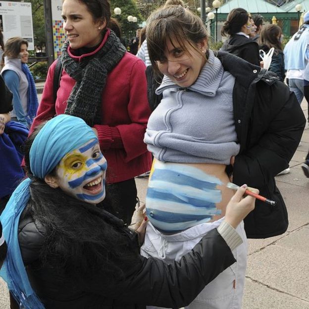 Mujer con la cara pintada con la bandera de Uruguay le dibuja la bandera en la panza a una embarazada.