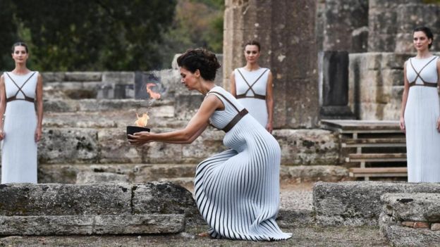 Atriz vestindo roupas helenísticas antigas acende a chama a partir dos raios de sol através de um espelho parabólico