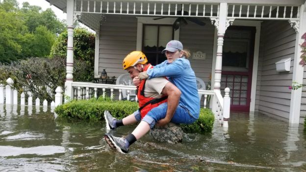 A soldier, waist-deep in water, carried a woman on his back past the front of a house porch