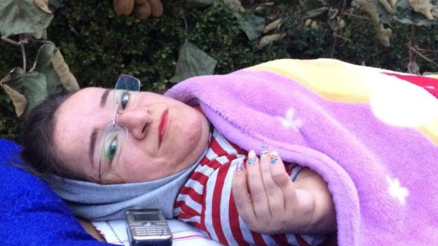 Mitra has a quilt wrapped around her and a mobile phone next to her. She is wearing spectacles in the picture and sporting nail polish.