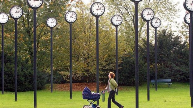 Una mujer camina con un cochecito de bebé en un parque con una instalación de muchos relojes