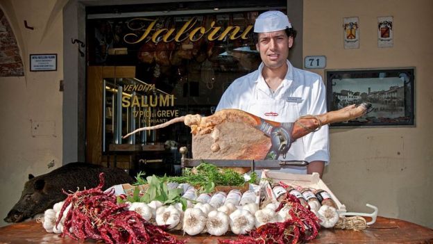 Italiano ofrece frente a su tienda una pata de jamón de cerdo.