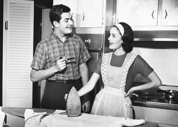Una pareja se mira en la cocina mientras ella sujeta la plancha y él una pipa.