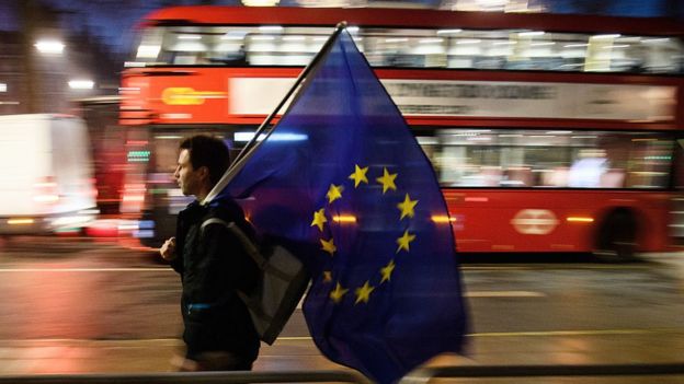 Joven con una bandera de la Unión Europea camina por la calle mientras atrás suyo pasa un bus double decker.