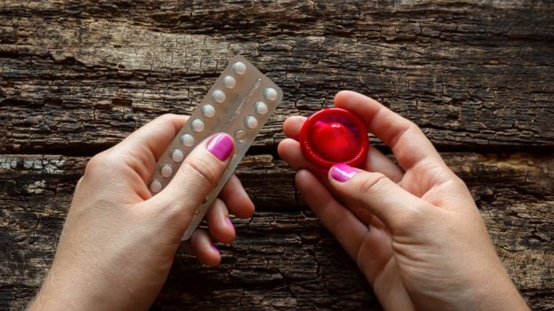 Mujer sostiene unas píldoras anticonceptivas con su mano izquierda y un preservativo con la derecha.