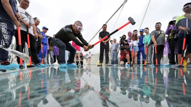 A man swings a sledgehammer at the floor of the glass bridge