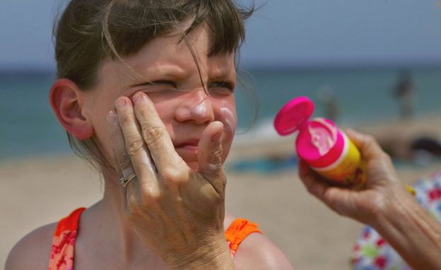 Mujer da crema en la playa a una niña.