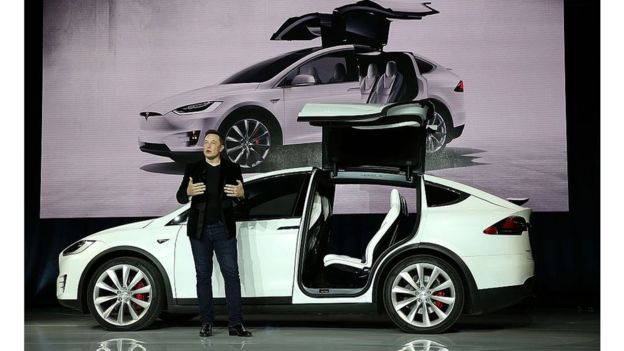 Elon Musk da una conferencia con un Tesla detrás