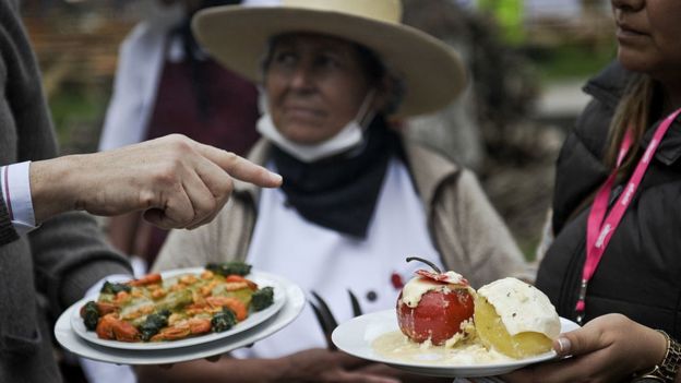 Mano de un hombre blanco y dos mujeres de origen andino frente a platos de comida.