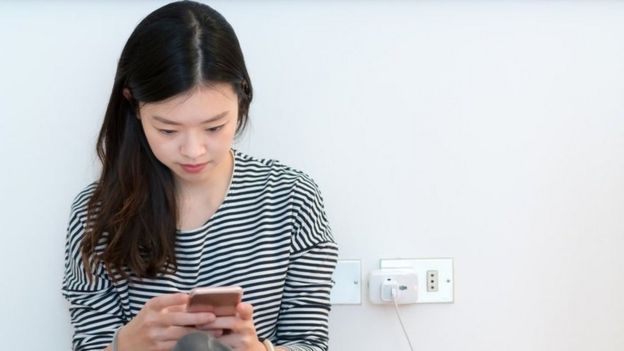 Joven cargando su celular en el enchufe de una pared.