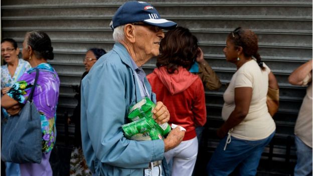 Un hombre pasa con su compra frente a una cola en Caracas