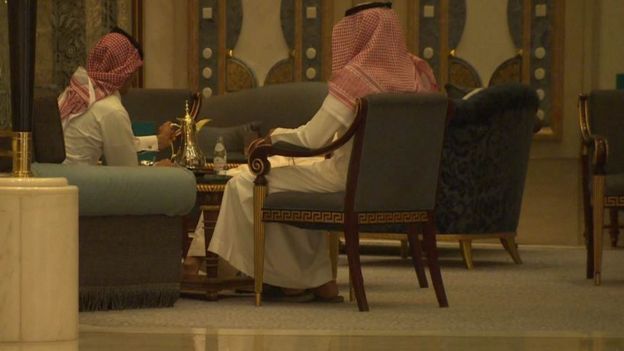 Saudimen in traditional dress sit in chairs in the Ritz Carlton in Riyadh, drinking coffee.
