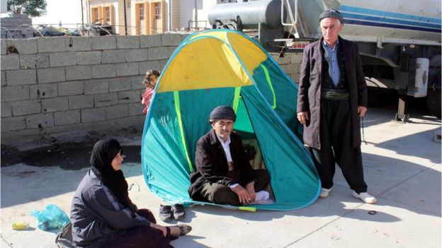 Man in doorway of tent pitched on street, with another man, a woman and a child visible around him