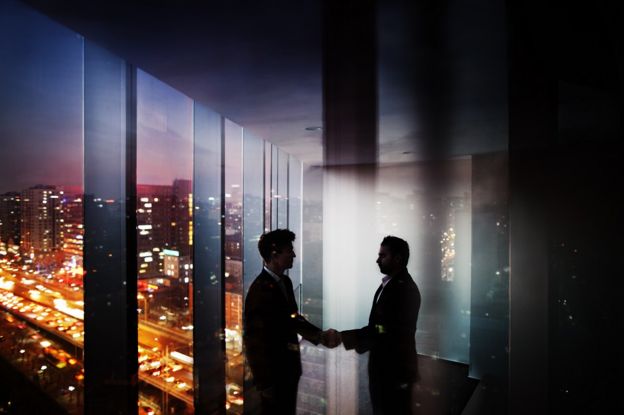 Two shadowy figures shake hands in a skyscraper
