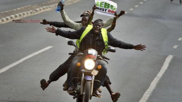 A motorcycle carrying protesters holding a placard using the acronym of the national electoral commission