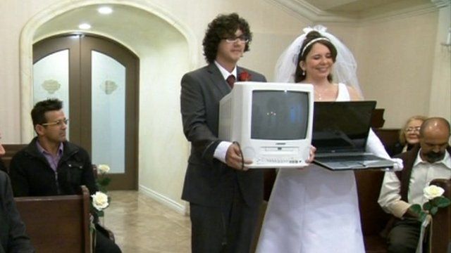 Staged "wedding" between man carrying a TV and a woman carrying a laptop.
