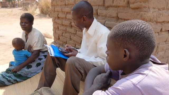 Woman with child being interviewed by a man holding a mobile phone and papers and a second man in the foreground