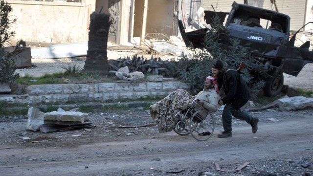 A civilian in a wheel chair is aided by an armed man ahead of being evacuated by United Nations (UN) staff, from the besieged district of the central Syrian city of Homs