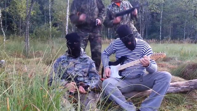 Masked men in military uniforms, one playing a guitar.