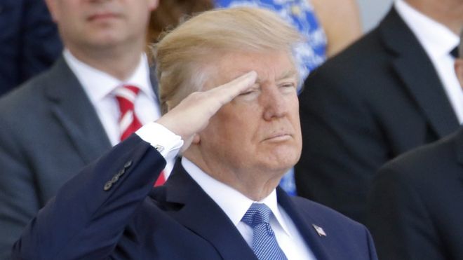 Donald Trump salutes, standing, from a viewing platform with other dignitaries