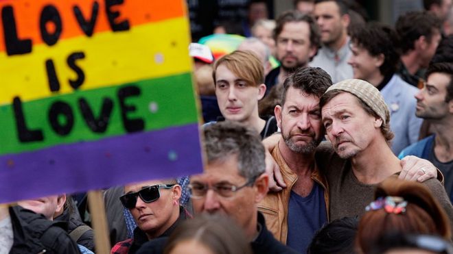 Δύο άνδρες αγκαλιάζουν πίσω από ένα σημάδι λέγοντας "η αγάπη είναι η αγάπη" σε ένα ράλι για την ισότητα των γάμων στην Αυστραλία
