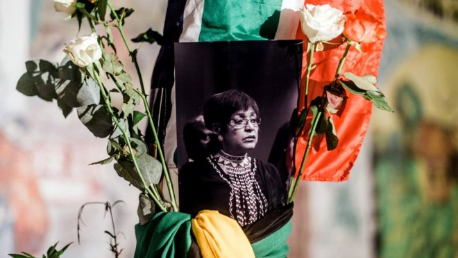 A black and white photograph of the late Winnie Madikizela-Mandela is surrounded by the South African and African National Congress (ANC) flags on a pole at the Old Durban Prison's Human Rights wall as South Africans gather to pay respect to the late high-profile anti-apartheid activist during a candle vigil in Durban on April 2, 2018.